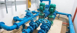 Closely arranged blue industrial pipes with handwheels and green motor-driven pumps in a utility room setting.