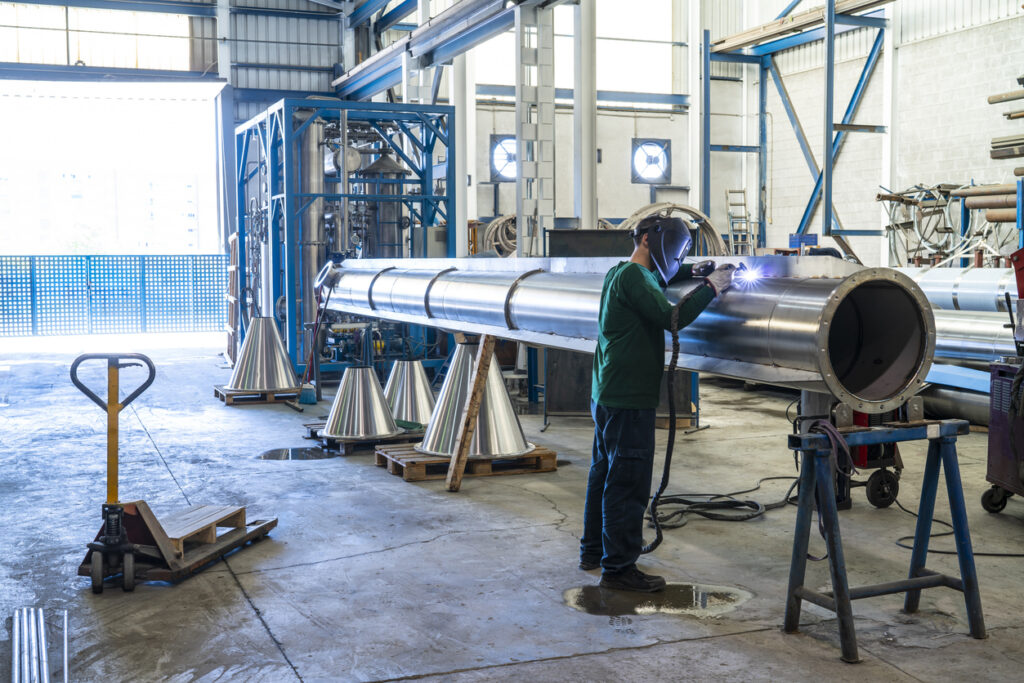 Welder working on steel pipe in industrial workshop with safety gear.